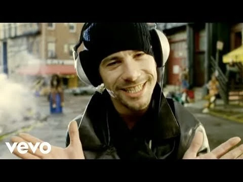 Smiling man wearing a black beanie, headphones, and a dark jacket gesturing in an urban street setting.