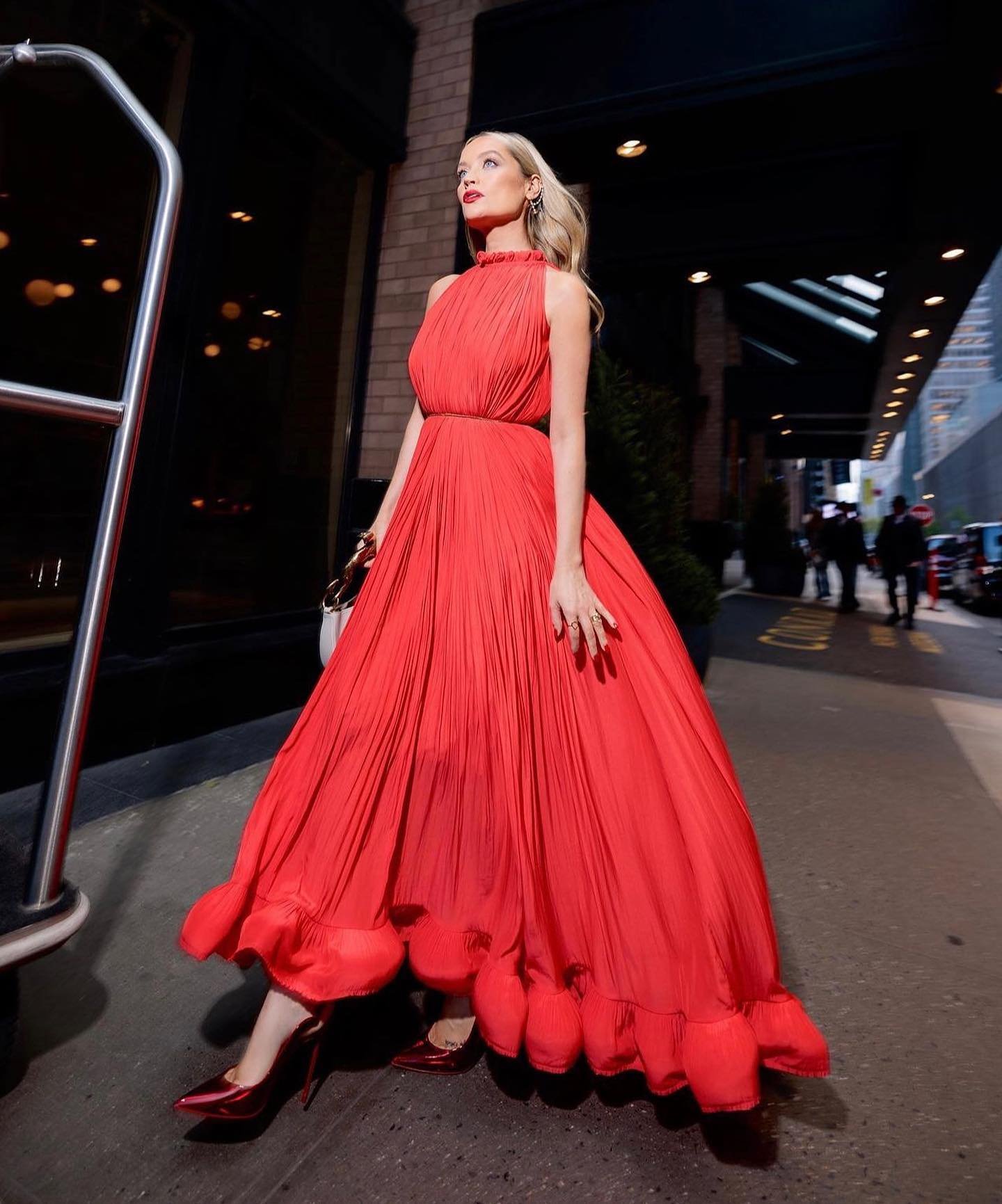 Woman in a flowing red dress stands outdoors on a city street with a building behind her, showcasing a blend of fashion and urban style.