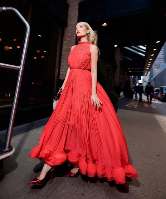 Woman in a flowing red dress stands outdoors on a city street with a building behind her, showcasing a blend of fashion and urban style.