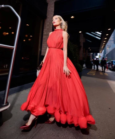 Woman in a flowing red dress stands outdoors on a city street with a building behind her, showcasing a blend of fashion and urban style.