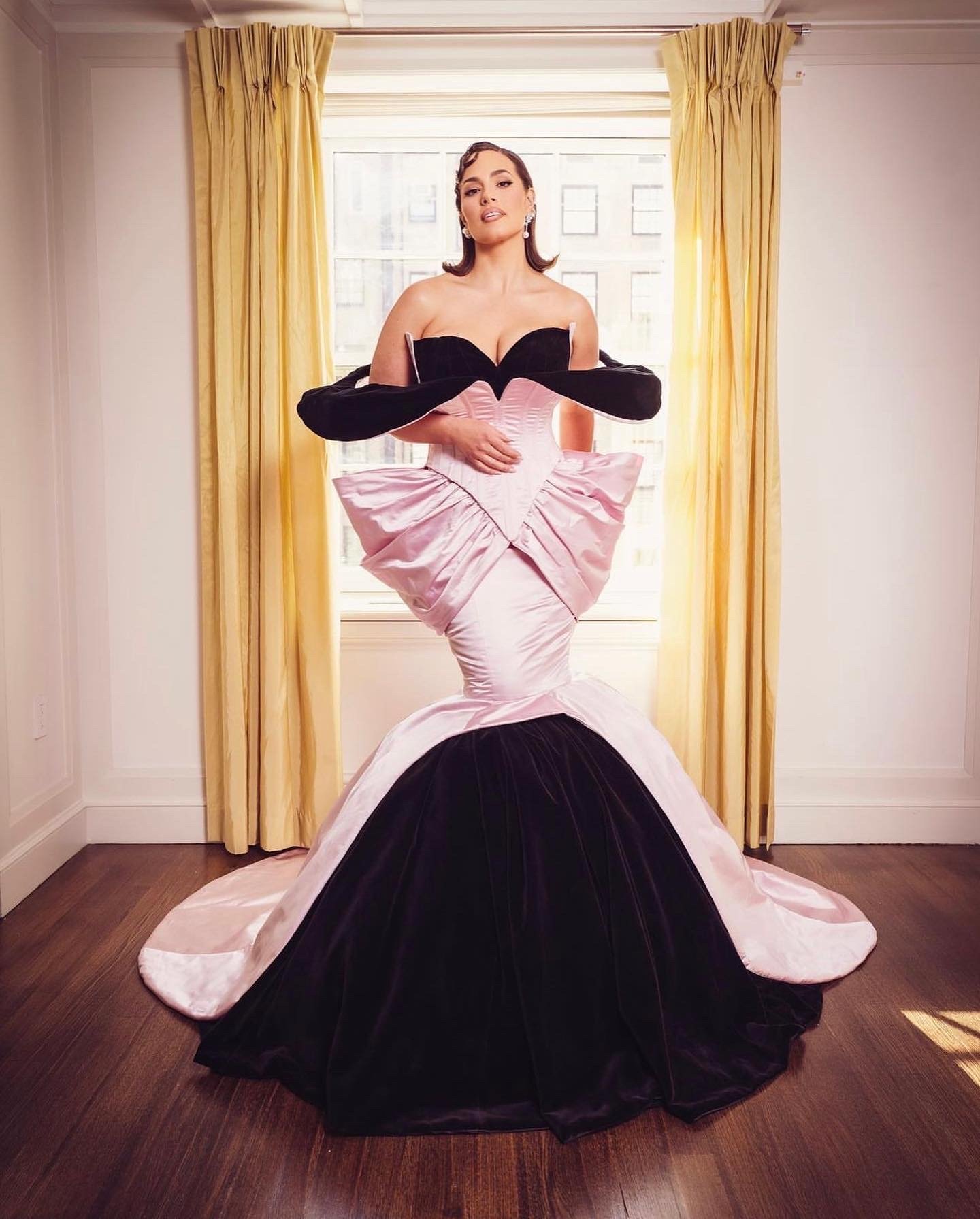 Ashley Graham poses in a black and pink strapless dress with a large bow detail in front of a window with golden curtains.