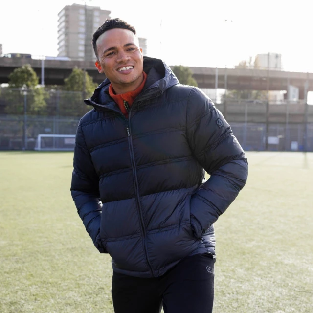 Smiling man in a hooded puffer jacket stands on a green sports field with city buildings in the background on a sunny day.