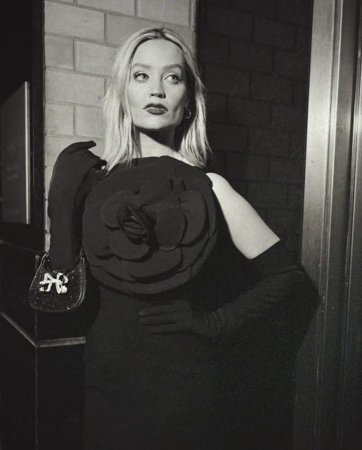 Black and white portrait of a fair-skinned woman in a dark dress with a large rosette, holding a small purse with a brick wall backdrop.