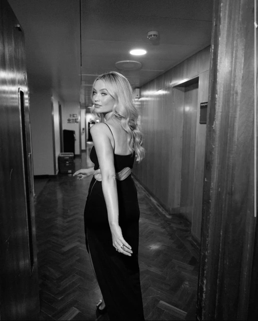 A blonde woman in a black dress looks over her shoulder in a hallway with wood paneling and herringbone flooring in a black-and-white photograph.