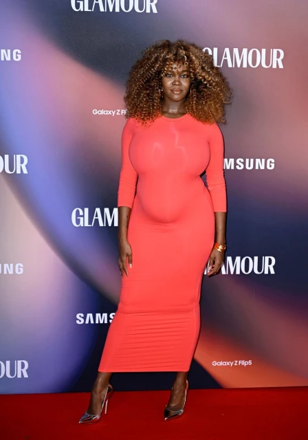 Adult woman with curly brown hair in a red dress and metallic high heels posing on a red carpet in front of a Glamour-branded backdrop.