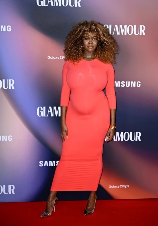 Woman with curly blonde hair poses in a coral-colored dress and silver heels against a branded backdrop on a red carpet indoors.