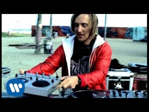 DJ mixing music at an outdoor event on a sound mixer, wearing a red jacket, with a beach and ocean in the background.