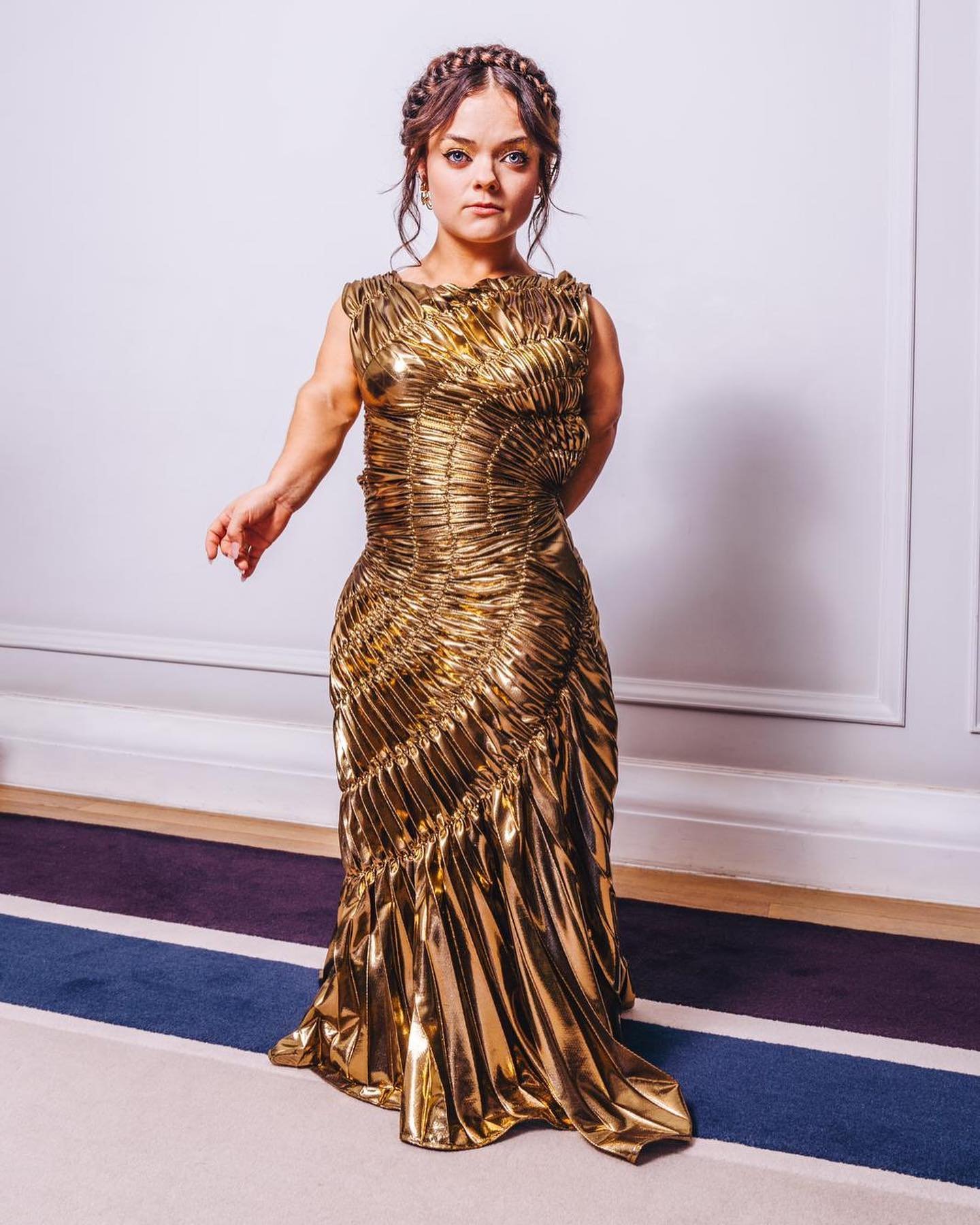 Woman in golden gown posing indoors. Female with braided hair wears a shiny, floor-length gold dress with a gathered bodice.