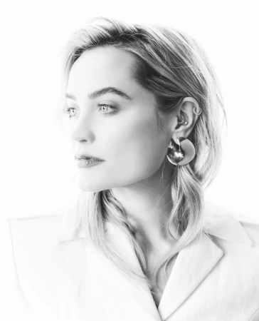 Close-up monochrome portrait of a woman wearing statement sculptural earrings and a collared shirt against a white backdrop.