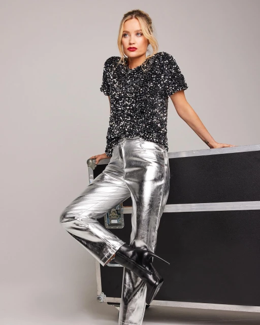 Woman in sequin top and silver pants poses next to black road case in a studio, showcasing a modern and edgy fashion aesthetic.