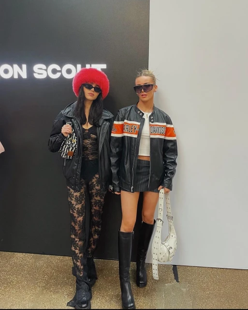 Two stylish adult women pose against a black and white wall, wearing matching leather jackets, boots, and sunglasses.