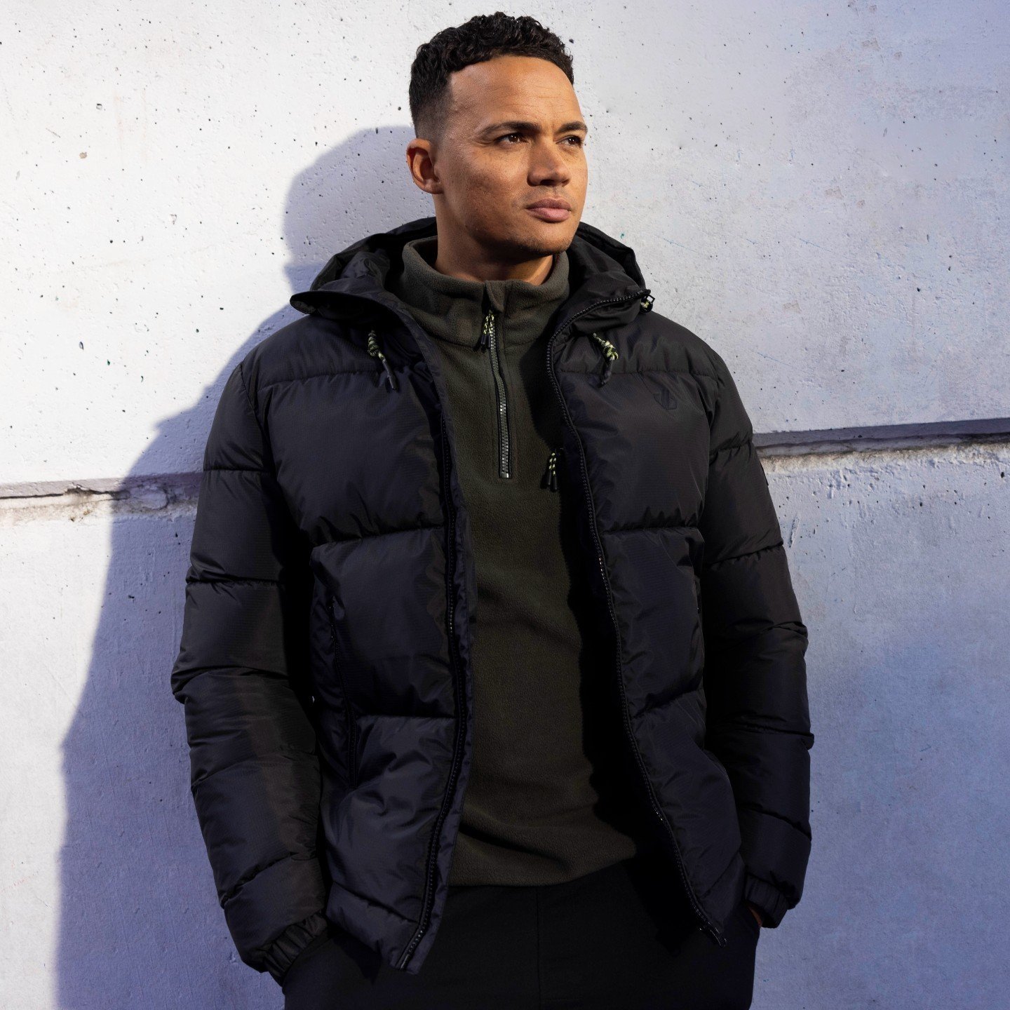 Adult male model posing outdoors with neutral expression wearing black puffer jacket, standing near textured gray wall in portrait orientation.