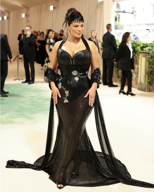 Ashley Graham poses on the red carpet wearing a full-length black gown with a train and floral embellishments at a fashion event.