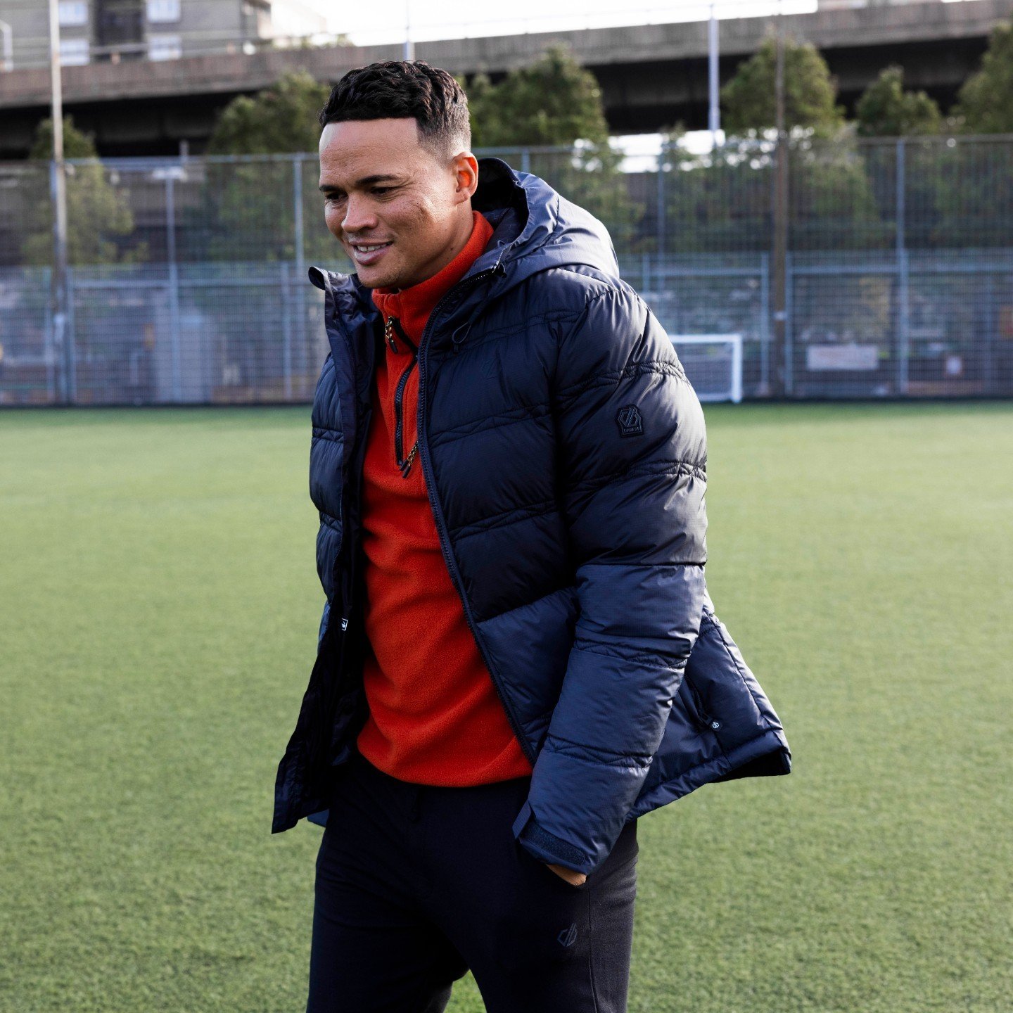 A man smiles on a soccer field, wearing a blue hooded puffer jacket and red fleece. He is on a green sports field with a chain-link fence.