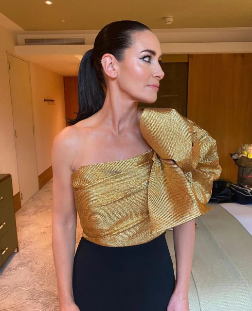 Adult woman poses in a black and gold one-shoulder dress with her dark hair pulled back indoors. Side profile shows her eye makeup.