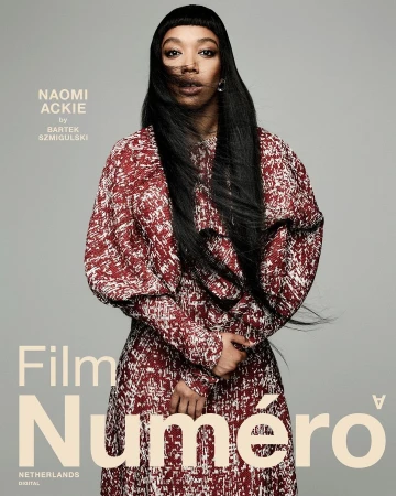 Portrait of Naomi Ackie wearing a red patterned long-sleeve dress against a plain grey background