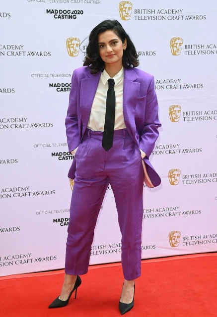 A woman in a purple suit on a red carpet smiles at the British Academy Television Craft Awards. She wears a black tie and heels.