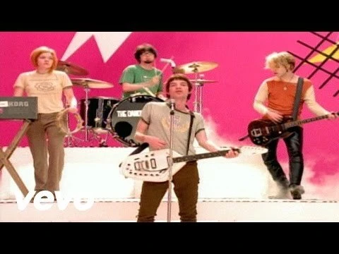 Band members perform on stage with instruments. Four people visible: drummer, keyboardist, guitarist and singer in front of pink backdrop.