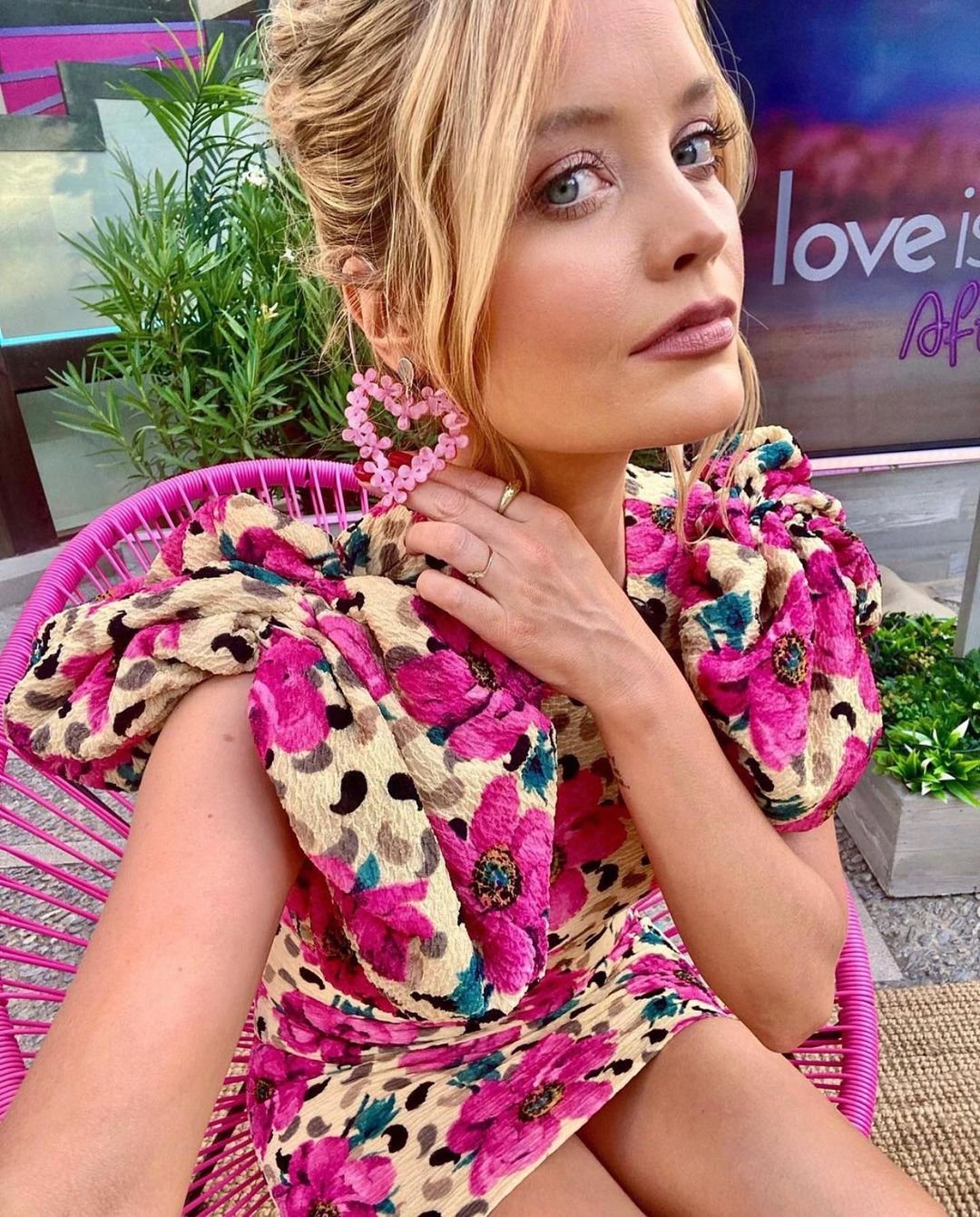 Close-up portrait of a woman with blonde hair in a pink floral dress and heart earrings, sitting in a pink chair outdoors.
