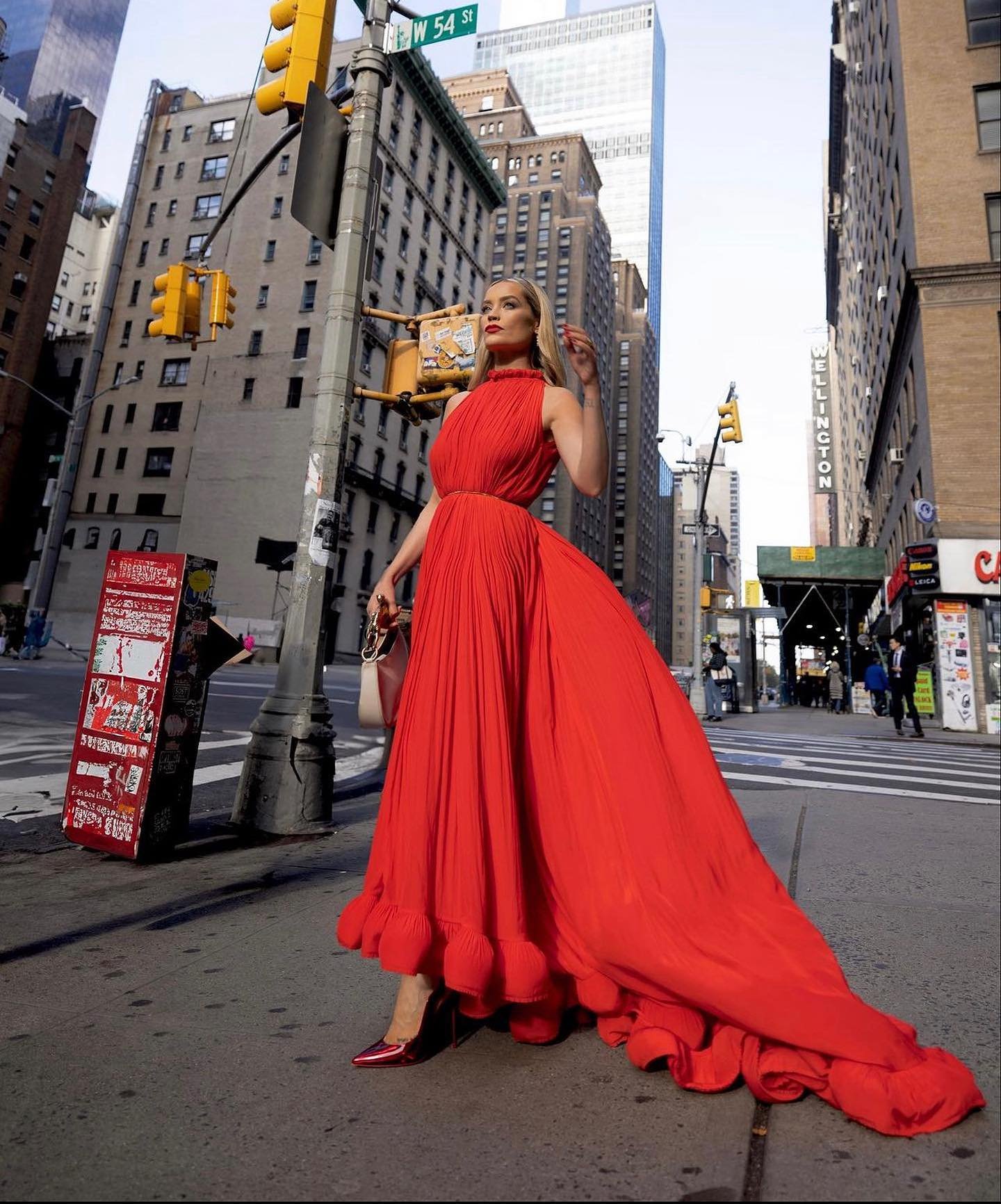 Woman in a long vibrant red pleated gown standing on a busy New York City street corner.