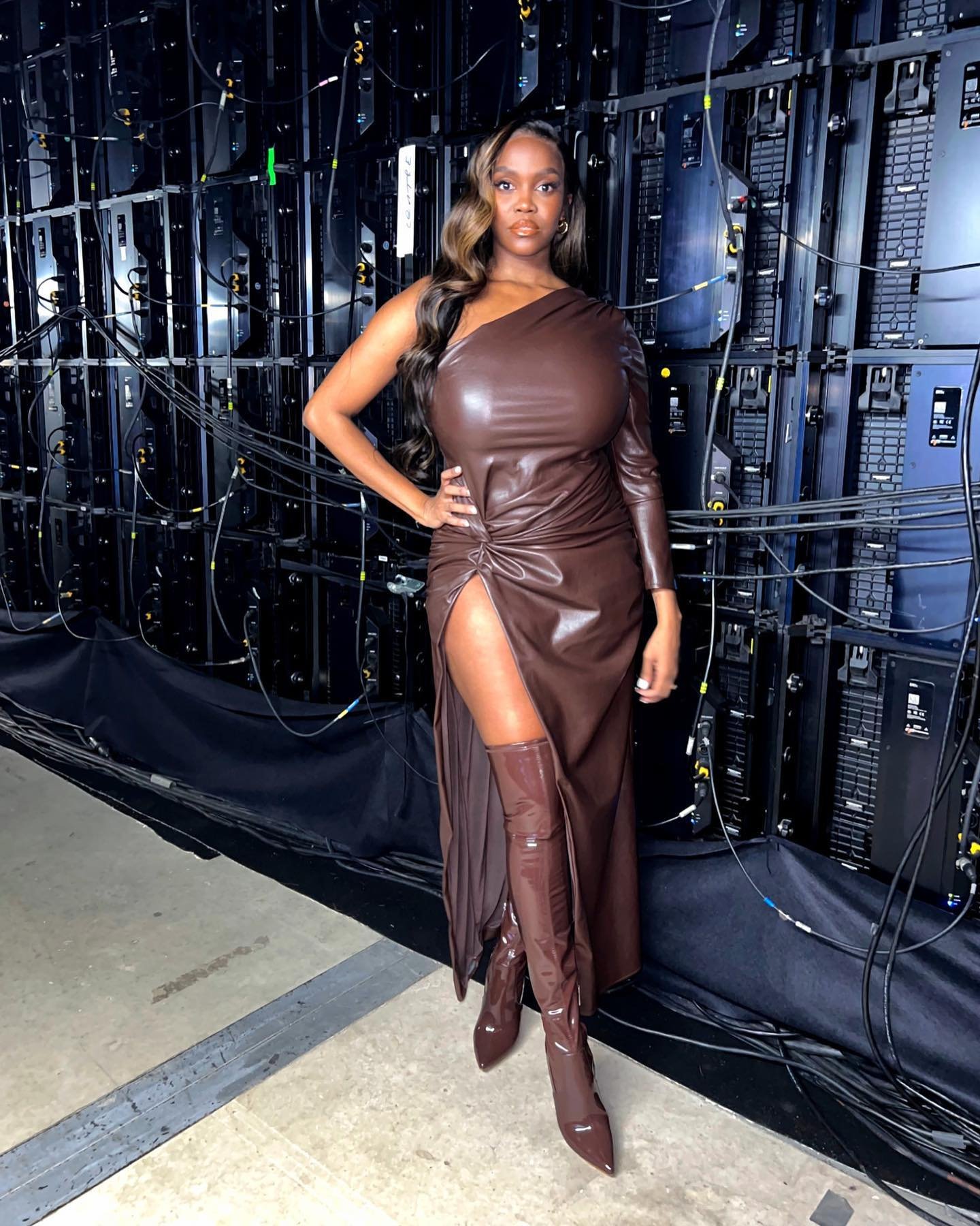 Attractive adult woman in a brown, one-shoulder dress and matching boots posing in front of a dark electronic equipment background.