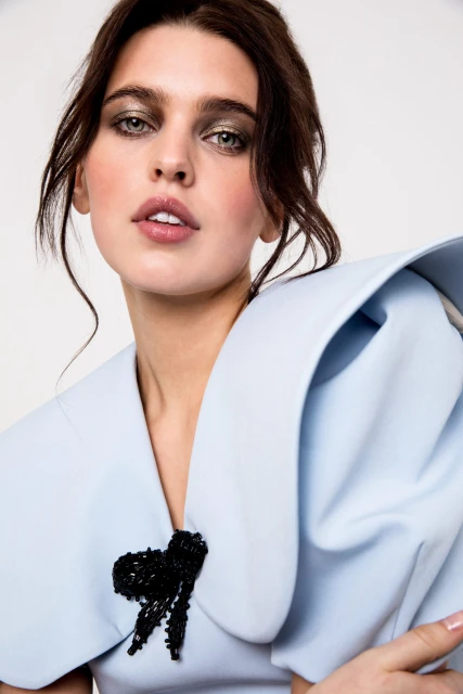 Headshot of a female fashion model wearing a light blue jacket with a black beaded bow, showcasing soft glam makeup in a studio setting.