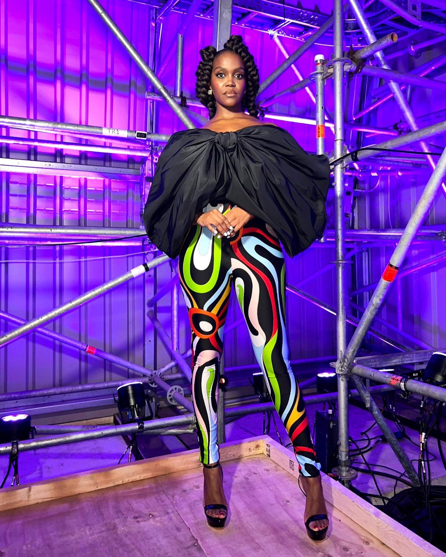 Elegant Black woman in black blouse with large bow and colourful leggings stands in front of a scaffolding and purple lighting.