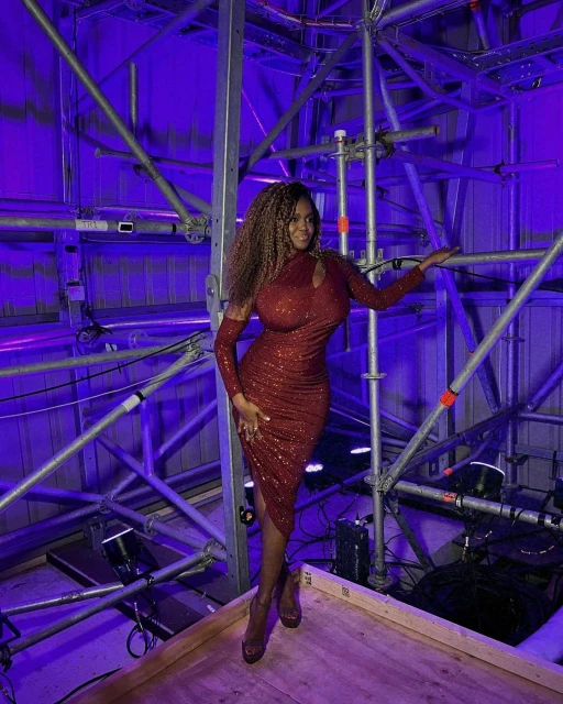 A woman with long wavy brown hair in a shimmering reddish brown dress and platform heels, posing against metal scaffolding lit with blue light.