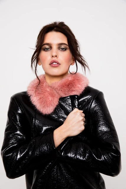 A fashionable woman poses indoors wearing a shiny black coat with a pink fur collar against a white backdrop in a studio setting.