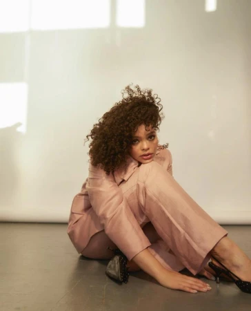 Woman with curly brown hair wearing a pink suit seated on a grey floor against a white wall.