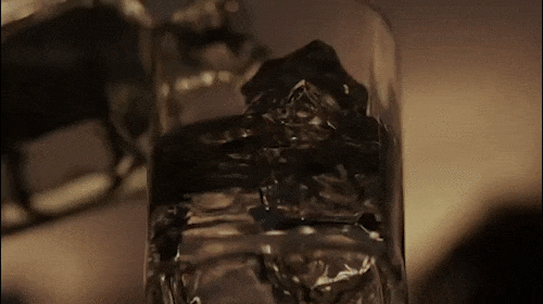Close-up of a clear glass filled with dark liquid and ice cubes, set against a neutral-toned background and softly lit.