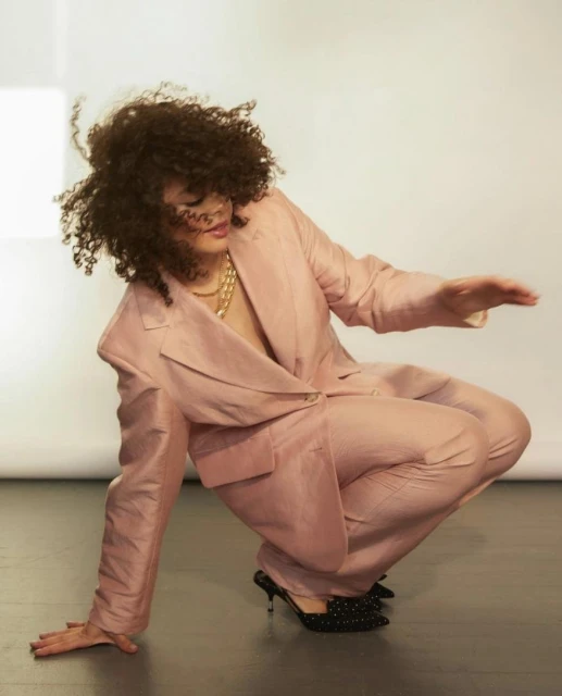 Woman in a blush-pink suit crouching indoors with curly hair, a gold chain, and black embellished mules against a neutral backdrop.