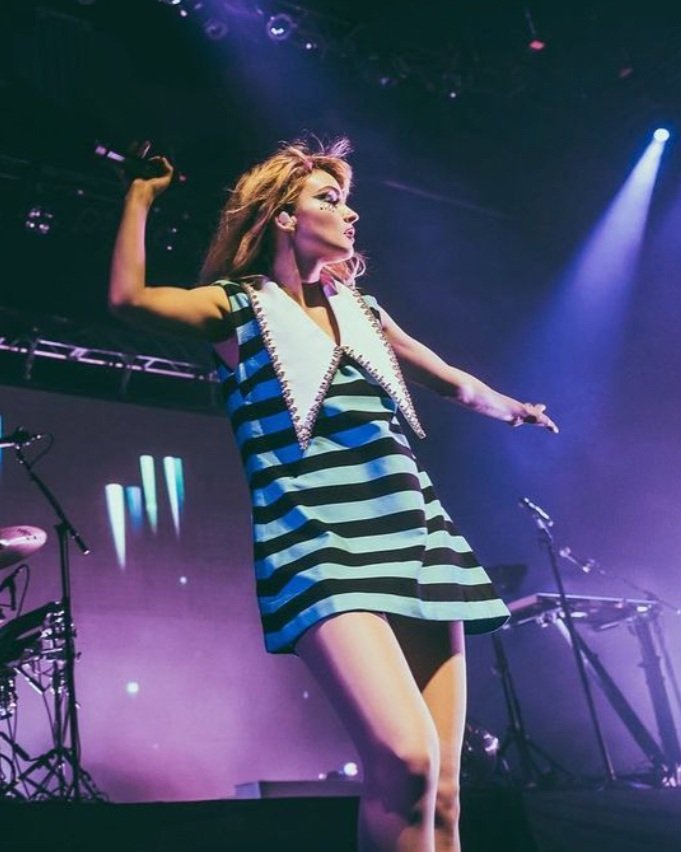 Female singer performing on stage with a microphone and wearing a retro-style mini dress with lights and musical equipment in the background.