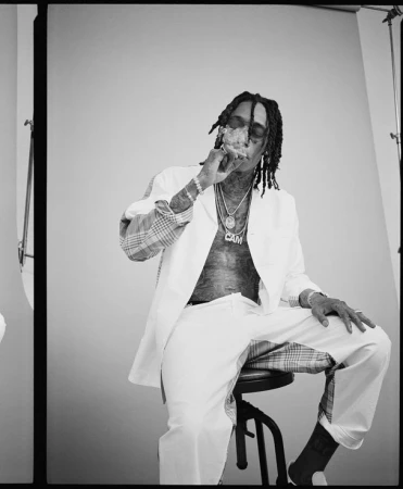 Black and white portrait of Wiz Khalifa seated on a stool, wearing a white shirt and trousers with patterned panels, smoking.