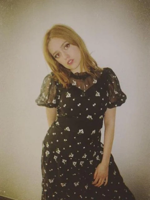 Young woman in a vintage-style floral dress poses indoors. The photo features a soft, faded aesthetic and shallow depth of field.
