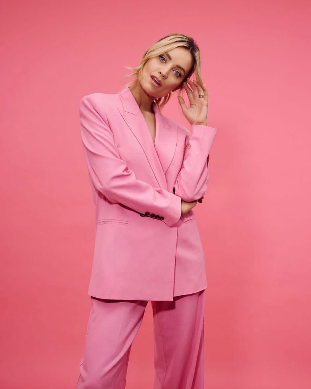 A woman in a pink blazer and pants poses in front of a pink background, looking at the camera with her hand near her head.