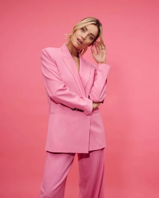 A woman in a pink blazer and pants poses in front of a pink background, looking at the camera with her hand near her head.