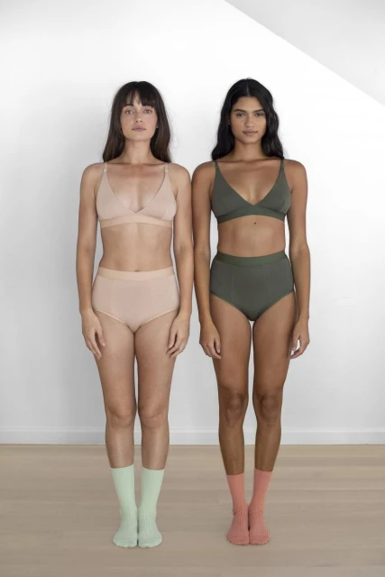 Two women stand side-by-side in underwear sets with colored socks against a white wall, showcasing fashion and style indoors.