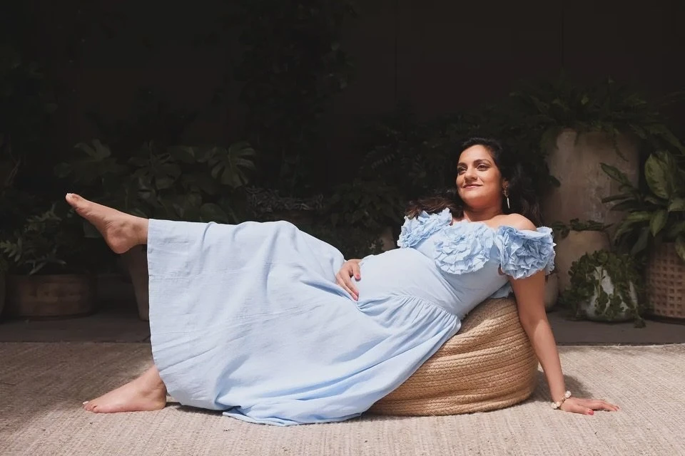 Pregnant woman in light blue dress reclines on a woven beanbag chair, surrounded by indoor plants against a dark background.