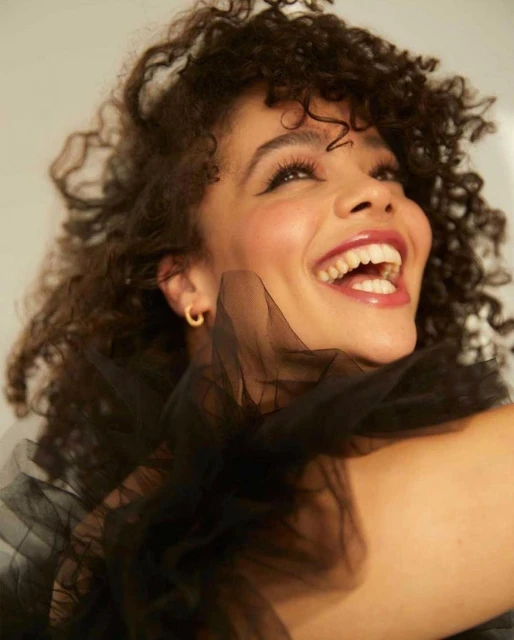 Portrait of an attractive young woman with curly hair laughing in a close-up shot, wearing a black tulle garment against a neutral background.