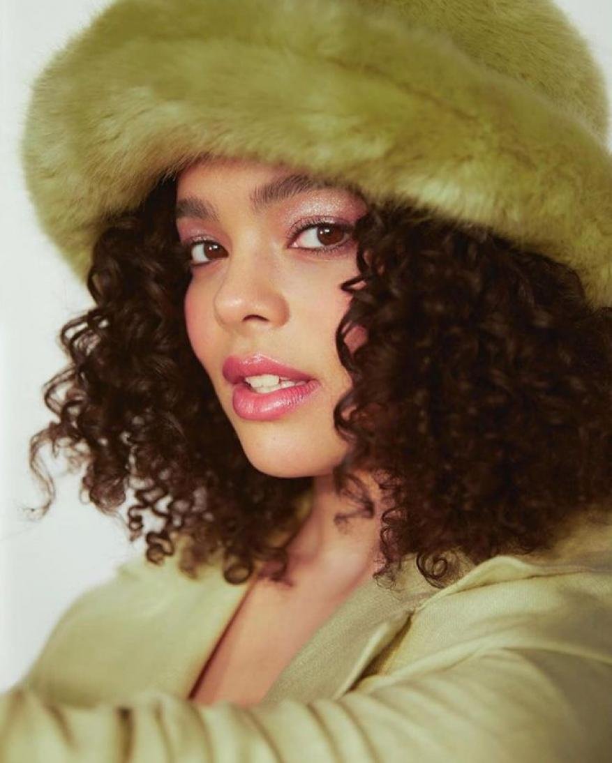 Portrait of an adult woman with curly hair wearing a green faux fur hat and matching green jacket against a plain white backdrop.