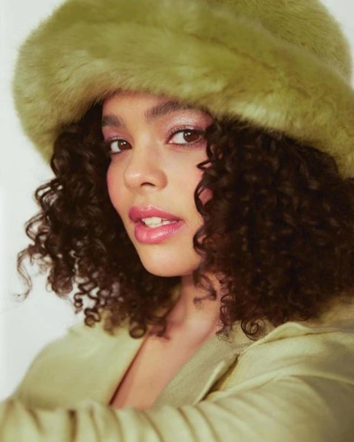 Portrait of an adult woman with curly hair wearing a green faux fur hat and matching green jacket against a plain white backdrop.