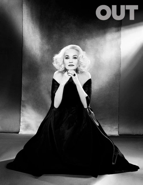 Black and white portrait of adult woman in long dress, fair skin, blond curly hair, indoor setting with 'OUT' logo in the upper-right corner.