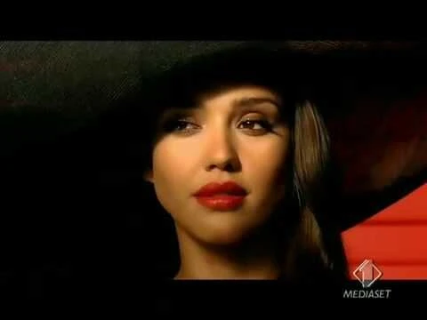 Close-up of a woman with red lipstick and tan skin in dark lighting. Woman gazes off-camera with a serious expression.