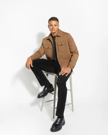 Man seated on a stool wearing a brown jacket, black trousers, and black loafers against a plain white studio background.