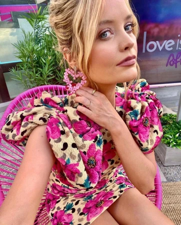 Blonde woman in floral dress wearing large pink heart earrings seated in a pink woven chair