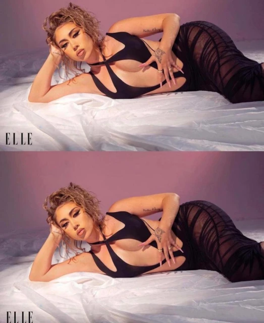 Female fashion model poses in a black cut-out dress on a white sheet in a studio setting with a pink background and soft lighting.