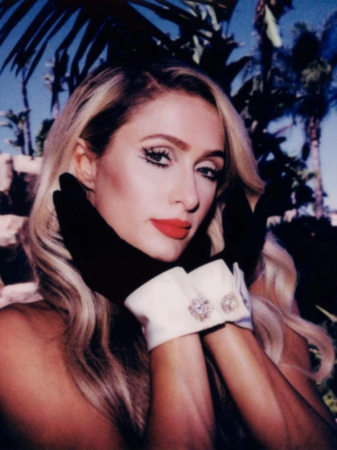 Close-up of a fair-skinned adult female with blonde hair, red lipstick, and striking eye makeup, wearing black gloves with jeweled white cuffs.