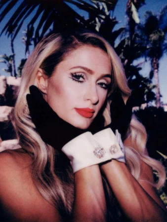 Close-up of a fair-skinned adult female with blonde hair, red lipstick, and striking eye makeup, wearing black gloves with jeweled white cuffs.