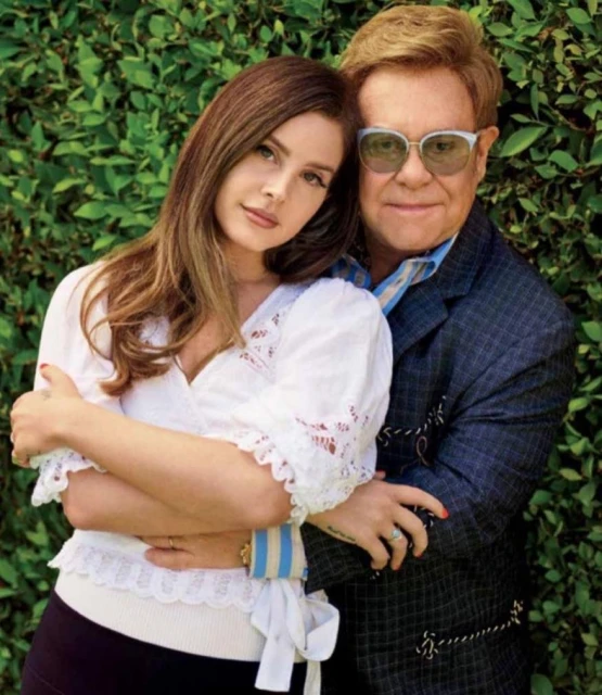 Singer Lana Del Rey poses with singer Elton John outdoors against a leafy green backdrop. They are closely posed and facing forward.